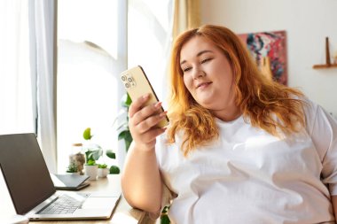 Işıl ışıl ve iri bir kadın ev içi yeşillik ve güneş ışığıyla çevrili akıllı telefonuyla meşgul olur..