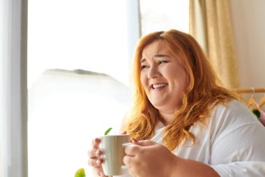 Neşeli, iri yarı bir kadın sıcak bir içkinin tadını çıkarıyor. Güneş ışığı penceresinden içeri süzülürken..