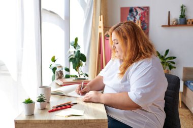 The beautiful plus size woman sits at a desk, drawing in a sketchbook, surrounded by plants. clipart