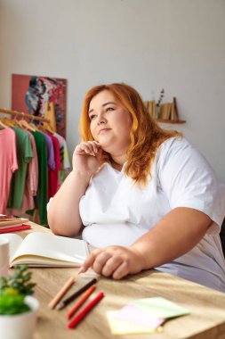 Büyük beden bir kadın, canlı kıyafetler ve sanat malzemeleriyle çevrili fikirleri düşünceli bir şekilde çizer..