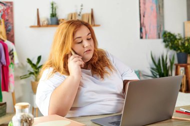 Kendine güvenen bir kadın telefonda konuşuyor, şık bir odada laptopuyla dikkatlice çalışıyor..