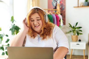 The plus size woman immerses herself in music, smiling while using her laptop in a lively room. clipart