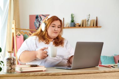 Kulaklıklı iri yarı bir kadın laptopuna odaklanırken kahvesini yudumluyor..
