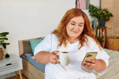 Neşeli ve iri yarı bir kadın dinlenme odasında telefonunu kontrol ederken gülümseyerek kahveyle rahatlar..