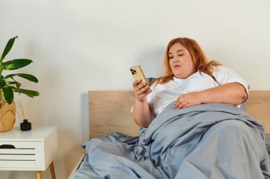 Büyük beden bir kadın yatakta dinleniyor, rahat battaniyelerin altında telefonuyla kayıyor..