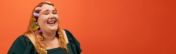 Радостный плюс размер женщина смеется, выставляя красочные заколки для волос у оранжевой стены.