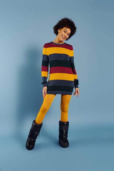Радостная молодая женщина в разноцветном полосатом свитере и желтых леггинсах светит в ярком пространстве.