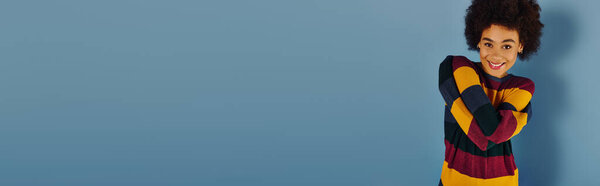 Молодая женщина в разноцветном свитере сияет радостью на мягком голубом фоне.