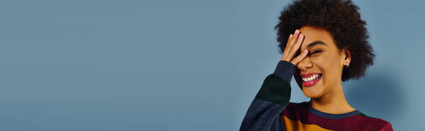 A joyful young woman smiles broadly, playfully hiding her face against a blue background.