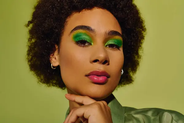 stock image A young woman displays an expressive look, adorned with striking green makeup and a colorful outfit.