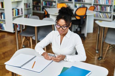 A dedicated teacher uses her smartphone while preparing lessons in a lively library environment. clipart