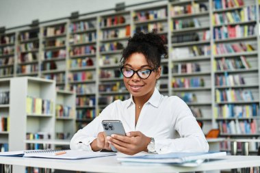 Heyecanlı bir Afrikalı Amerikalı öğretmen etrafı kitaplarla çevriliyken telefonunu kontrol ediyor..