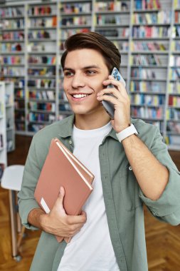 Genç bir adam kütüphanede telefonla konuşurken gülümsüyor ve kitapları tutuyor..