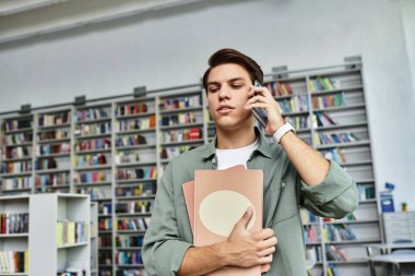 A dedicated student is studying intently on the phone while holding documents in a library. clipart