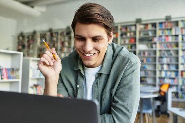 Neşeli bir genç adam, kitap raflarıyla çevrili, iyi aydınlatılmış bir kütüphanede özenle çalışıyor..