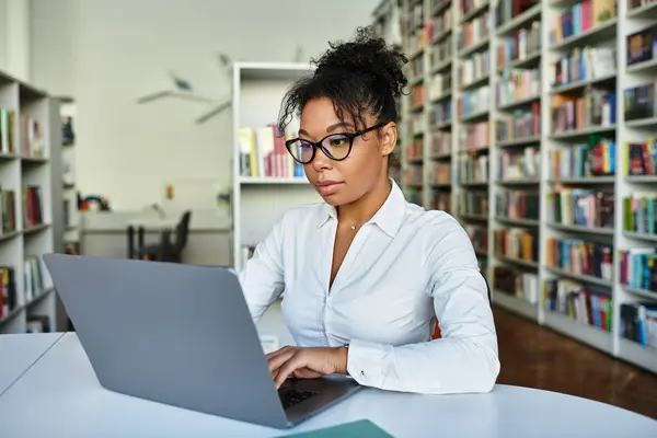 stock image A focused African American teacher utilizes a laptop in a vibrant library to enhance learning.