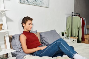 Kırmızı kolsuz tişörtlü genç bir kadın evinde rahat bir şekilde uzanıyor, huzurlu rutininin tadını çıkarıyor..