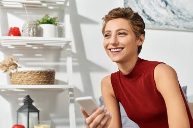 Kırmızı kolsuz tişörtlü neşeli genç bir kadın telefonuyla uğraşırken evde rahatlıyor ve neşe saçıyor..