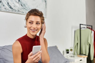 Kısa saçlı genç bir kadın evde dinleniyor, müzik dinliyor ve kırmızı kolsuz gömleğiyle gülümsüyor..