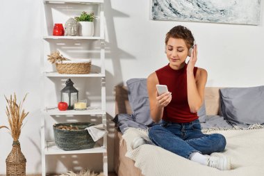 Kırmızı kolsuz tişörtlü genç bir kadın yatağında bacak bacak üstüne atmış bir şekilde oturuyor ve evinde dinleniyor..