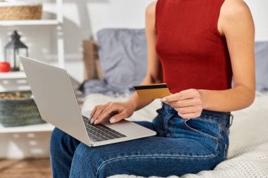 Kırmızı askılı bluz giyen genç bir kadın evde rahatça alışveriş yapıyor ve bilgisayarına odaklanıyor..