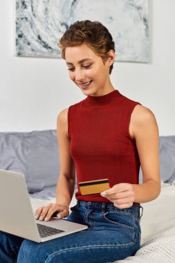 Kırmızı kolsuz tişörtlü genç kadın evde kredi kartıyla online alışveriş yapıyor..