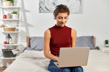 Sıcak bir yatak odasında, kırmızı atletli genç bir kadın laptopunu kulaklıkla kullanıyor, görevlerine odaklanıyor..