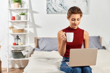 Kısa saçlı genç bir kadın evde rahatlıyor, kahvesini yudumluyor ve rahat bir ortamda laptopuna odaklanıyor..