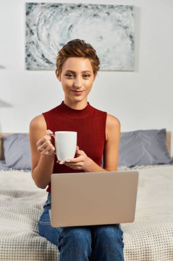 Yatakta rahat bir şekilde oturan genç bir kadın laptopuyla çalışırken kahvesini yudumluyor ve rahatlıyor..