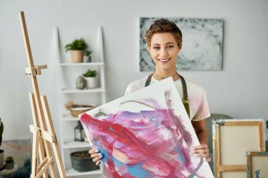 A young woman, sporting short hair and natural makeup, happily showing her painting in her inviting home studio space. clipart