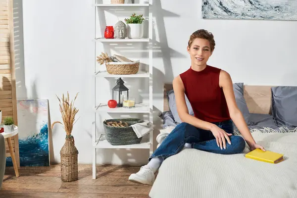 stock image In a serene living space, a young woman relaxes in a red tank top, reading a book with a calm demeanor.