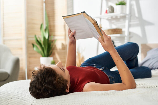 Relaxation and tranquility fill the air as a young woman reads a book on her bed in natural light.