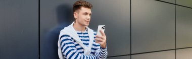 Protez bacaklı şık bir genç adam telefonuna gömülmüş, parlak bir duvara yaslanıyor..