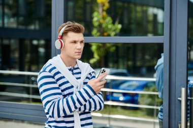 Yakışıklı bir genç adam dışarıda beklerken akıllı telefonuyla meşgul olarak müzik dinler..