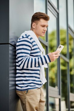 A young man with a prosthetic leg leans against a modern building, focused on his phone. clipart