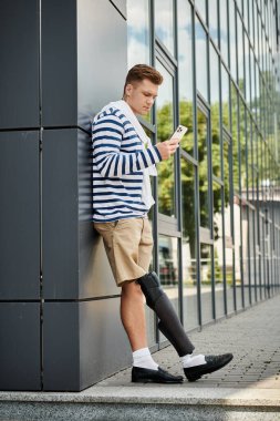 Yakışıklı genç bir adam, telefonuna odaklanmış, şık, modern bir binaya karşı duruyor..