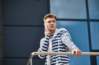 A young man with a prosthetic leg stands on a balcony, chatting on the phone under clear skies. clipart