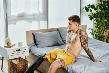 Protez bacaklı düşünceli genç bir adam bir yatağa oturur ve ışık dolu odaya bakar..