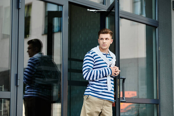 Молодой человек в полосатом свитере и хаки задумчиво смотрит за стеклянную дверь.
