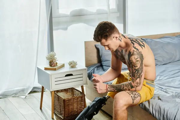 stock image A young man sits thoughtfully by his bed, showcasing his tattoos and prosthetic leg.