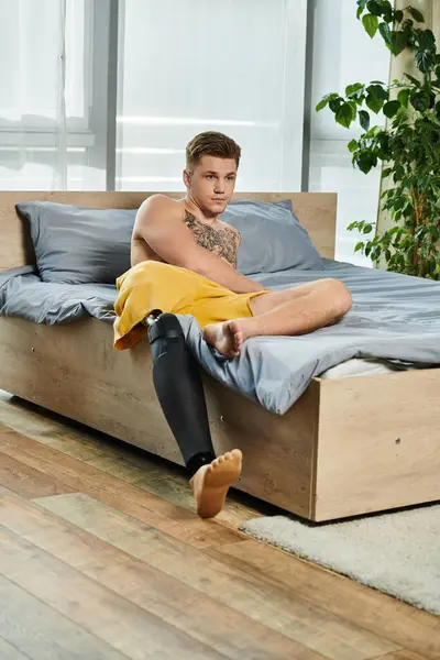 stock image A young, handsome man sits on his bed, showcasing his prosthetic leg with confidence and pride.