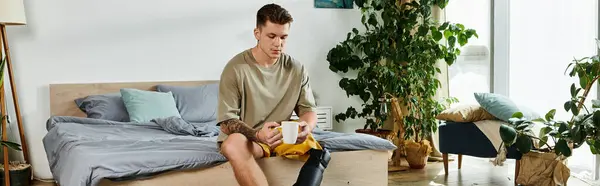 stock image A young man with a prosthetic leg sits on a bed, reflecting in a sunlit, plant filled room.