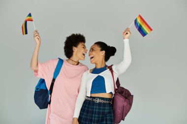 İki arkadaş sevinçle gökkuşağı bayrakları sallıyor, parlak bir atmosferde gurur ve birlikteliklerini kutluyorlar..