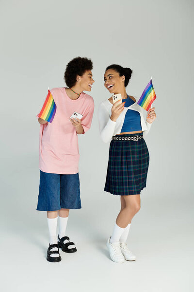Two friends joyfully share a moment while showcasing pride flags and engaging with their phones.