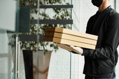 Siyah maskeli bir teslimatçı öğle yemeği saatlerinde modern bir ofis binasına çoklu pizza kutuları getirir..
