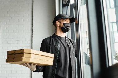 Siyah tıbbi maske takan bir kurye, modern bir ofis binasına birden fazla yiyecek kutusu taşır..