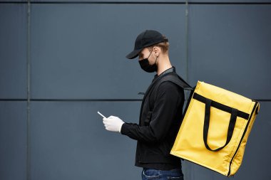 A delivery man wearing a black mask and gloves checks order details while standing by an office building. clipart