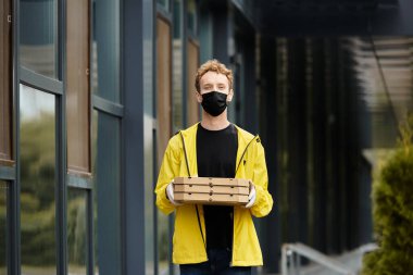 Siyah maskeli bir teslimatçı yemek için birkaç pizza kutusu taşıyan bir ofis binasına gelir..