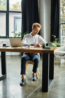 Kızıl saçlı odaklanmış bir işadamı, çağdaş ofis çalışma alanındaki bir proje üzerinde gayretle çalışıyor..