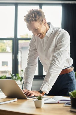 Beyaz tişörtlü, kızıl saçlı bir işadamı, şık ve modern bir ofis alanında dizüstü bilgisayarla çalışıyor..